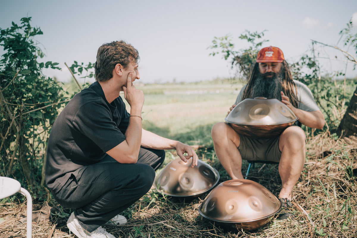 Two men in a grassy field with handpans, one playing a handpan on his lap while the other listens